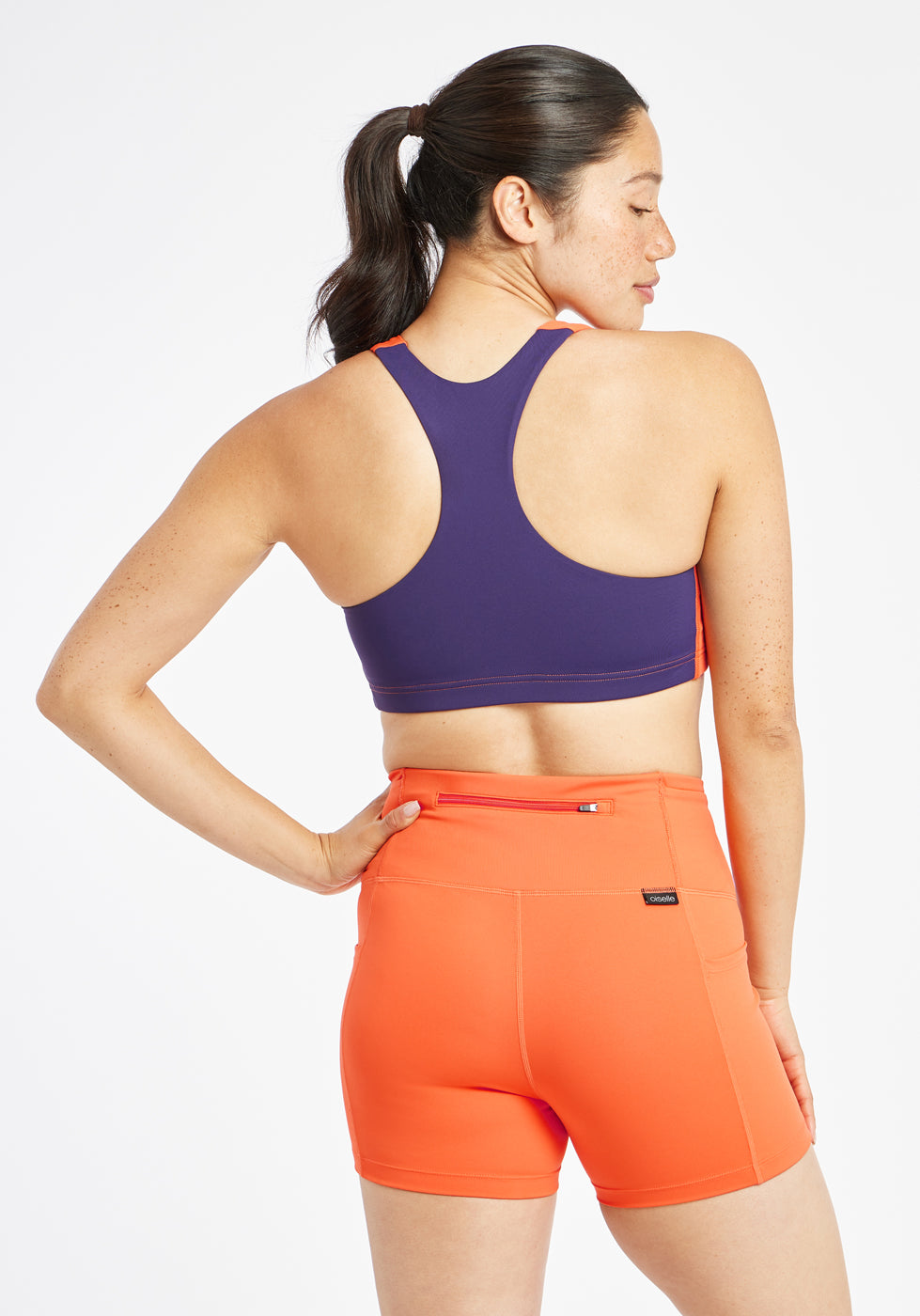 Woman wearing orange and purple paneled sports bra, alternate back view