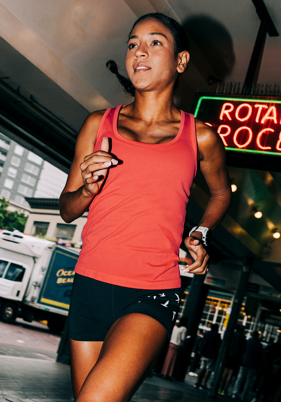 Woman wearing a red orange running tank top and black compression shorts, running through Pike Place Market