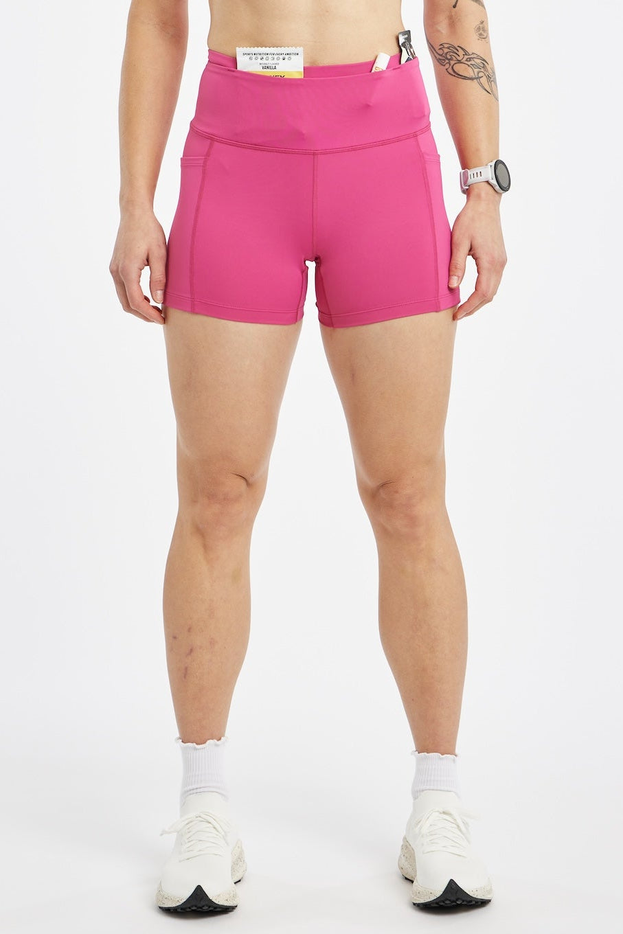 A woman modeling a bright pink athletic set consisting of a sports bra and matching high-waisted shorts, standing against a plain light gray studio background.