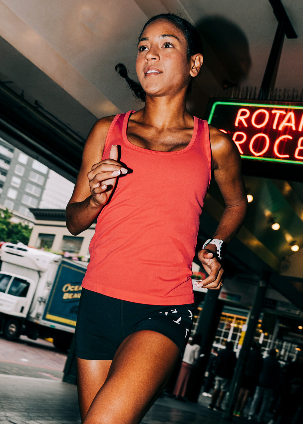 Woman wearing a red orange running tank top and black compression shorts, running through Pike Place Market