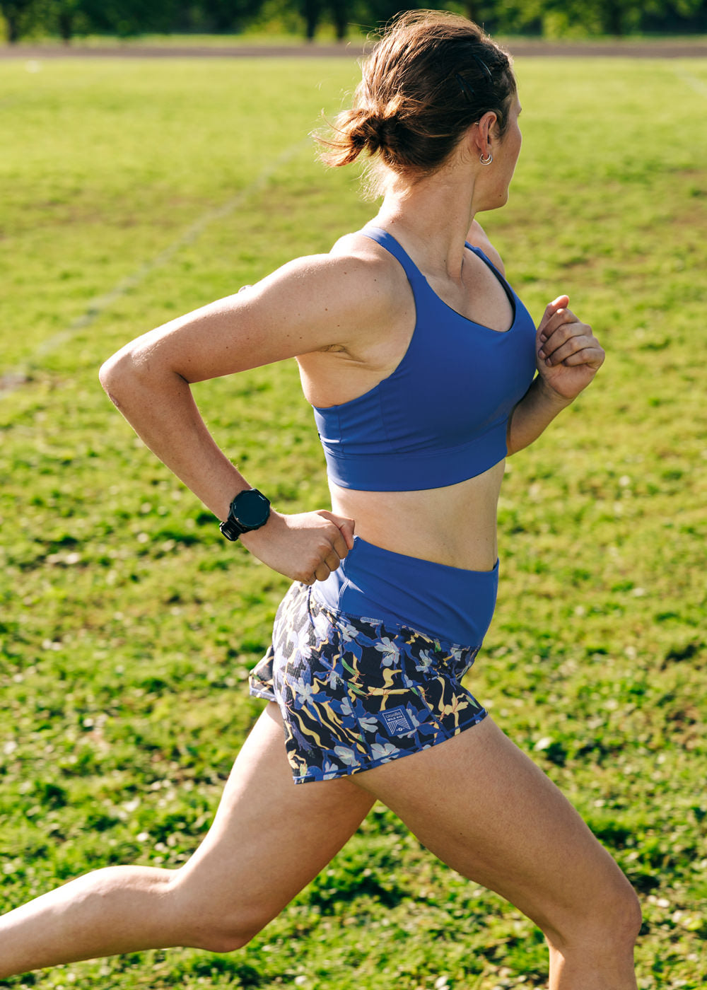 Woman running on a track in High Rise Roga 4" Running Shorts in blue floral print and matching blue Oiselle bra
